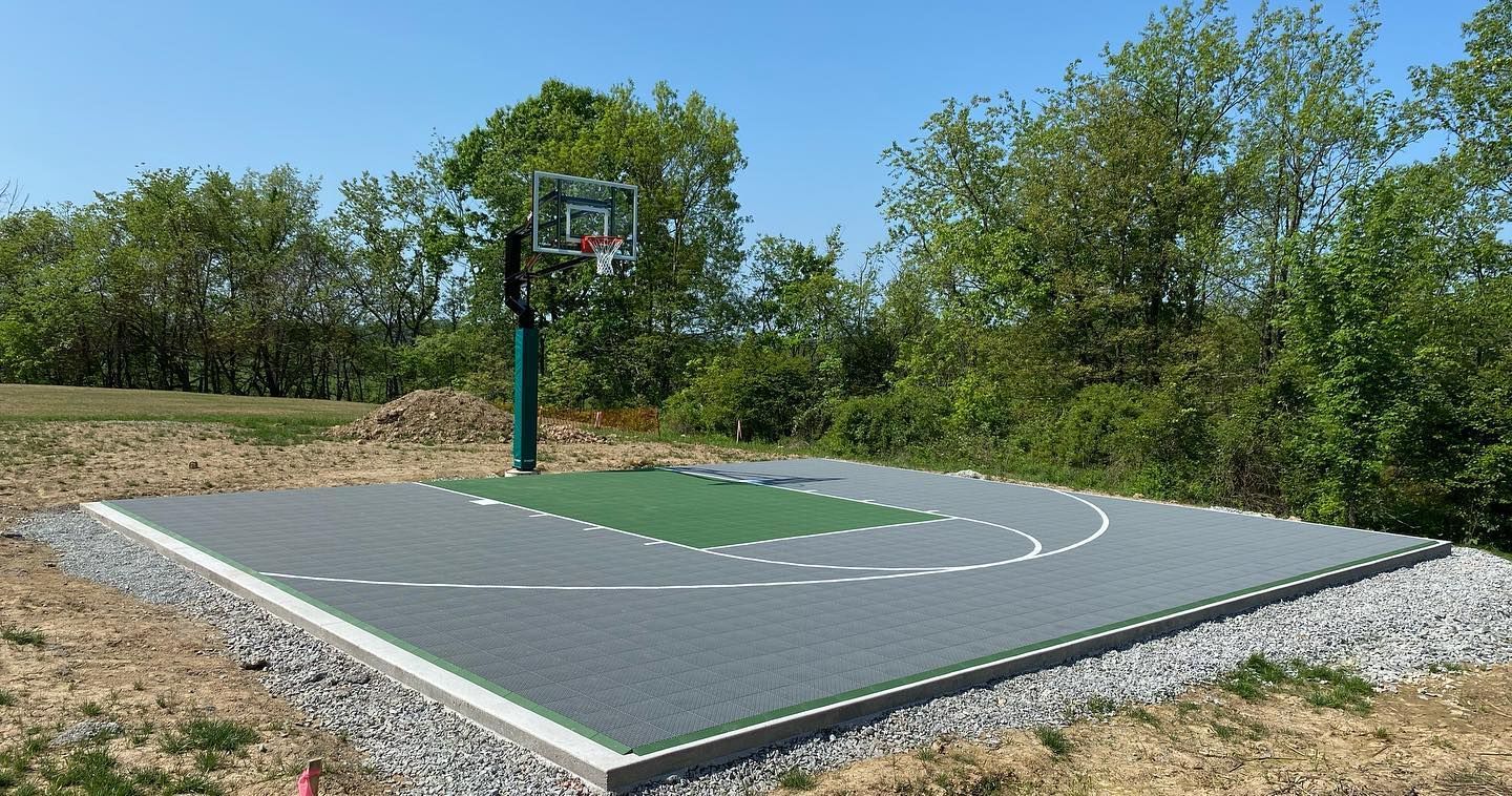 Outdoor basketball half-court with a hoop, gray surface, and white lines surrounded by trees.