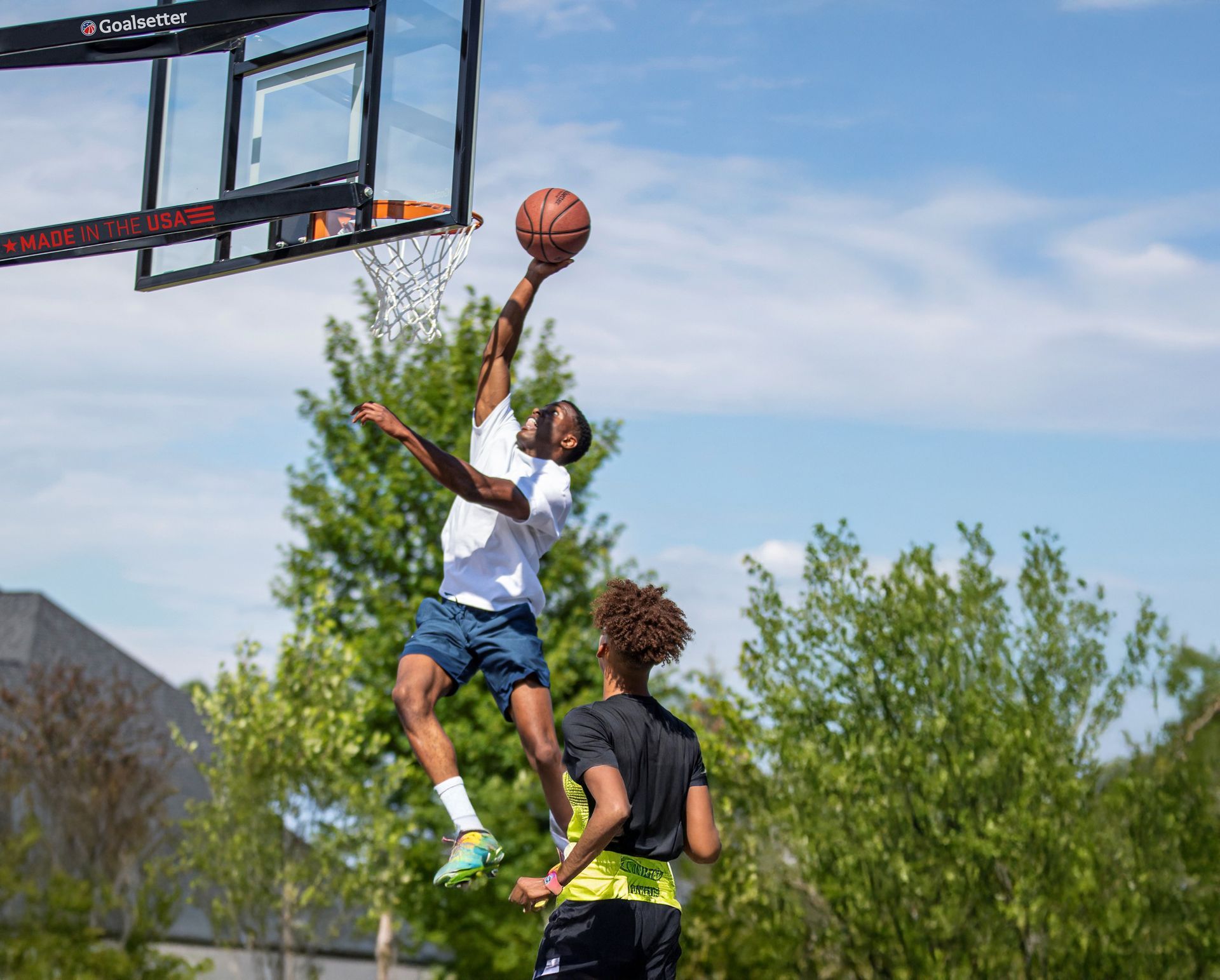 Basketball player in white jersey leaps for a layup while another defends outdoors near a hoop.