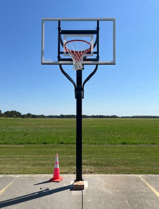 Outdoor basketball hoop on a court with a traffic cone beside it and open fields in the background.