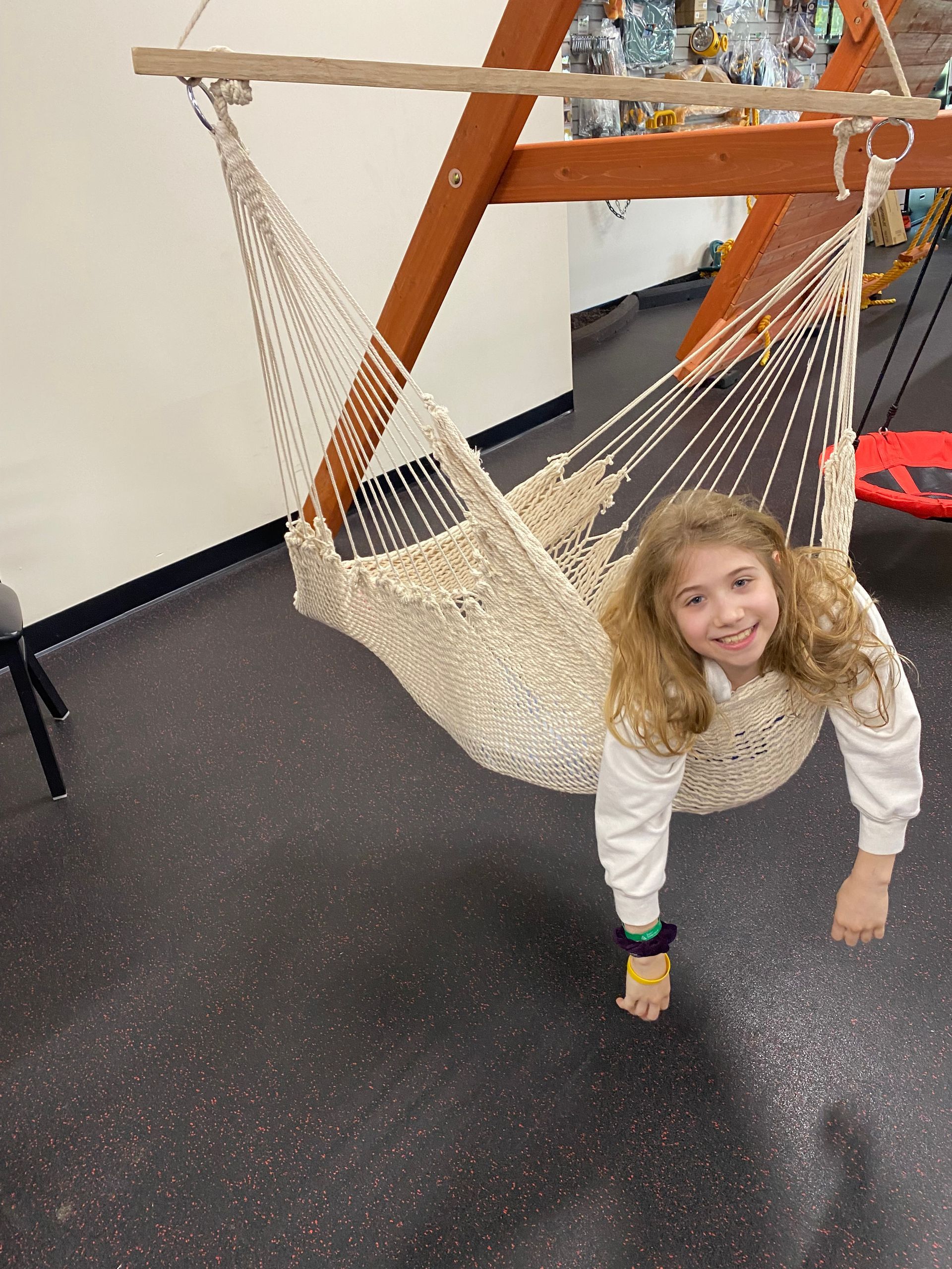 Child smiling in a woven hammock swing indoors, with orange frame and dark speckled floor.