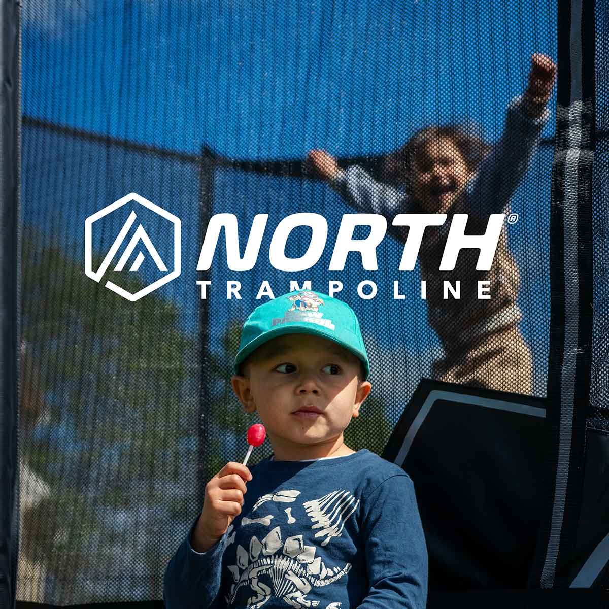 Child holding a lollipop in front of a North Trampoline net, with a person bouncing in the background