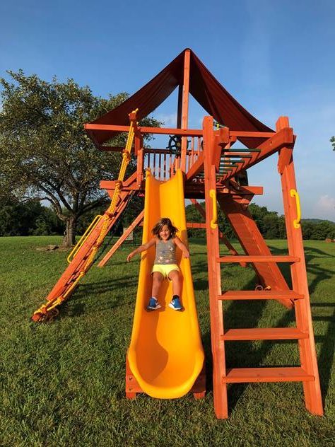 Child on yellow slide at a red wooden playground in a grassy park, with trees in the background