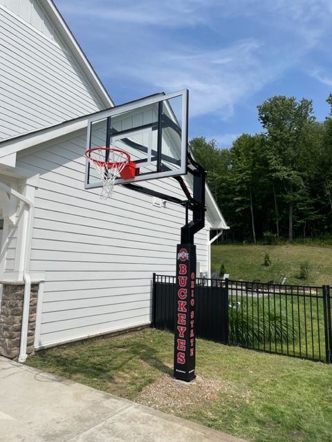Basketball hoop mounted beside a white house and black fence in a grassy yard.