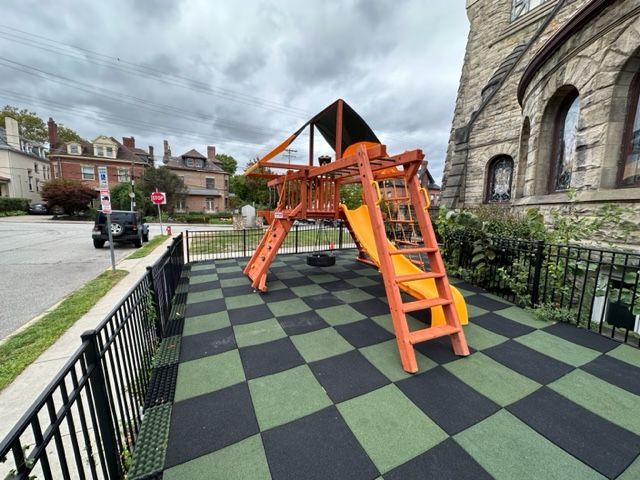 Orange playground climbing structure on a black-and-green checkerboard play surface beside a stone building.