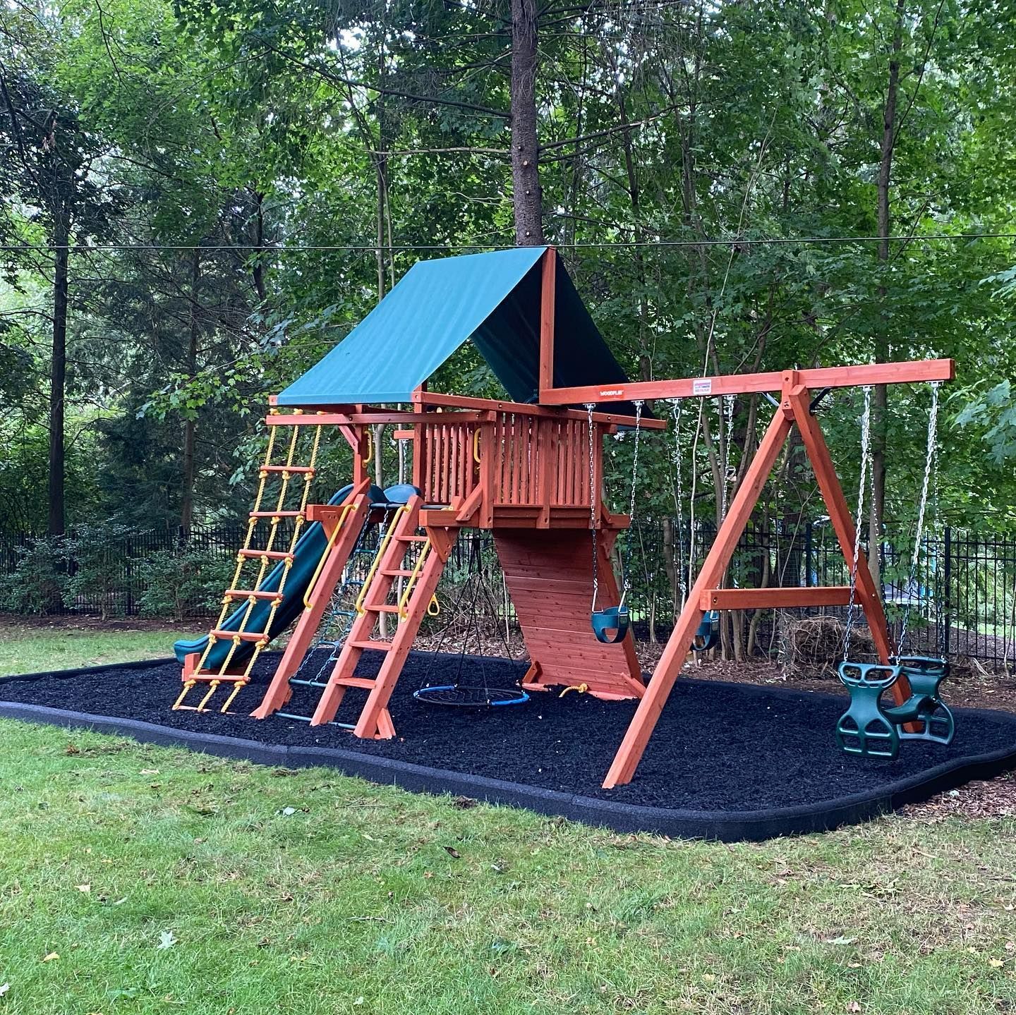 Wooden backyard playground with blue roof, slides, ladder, swings, and tree-shaded rubber mat ground
