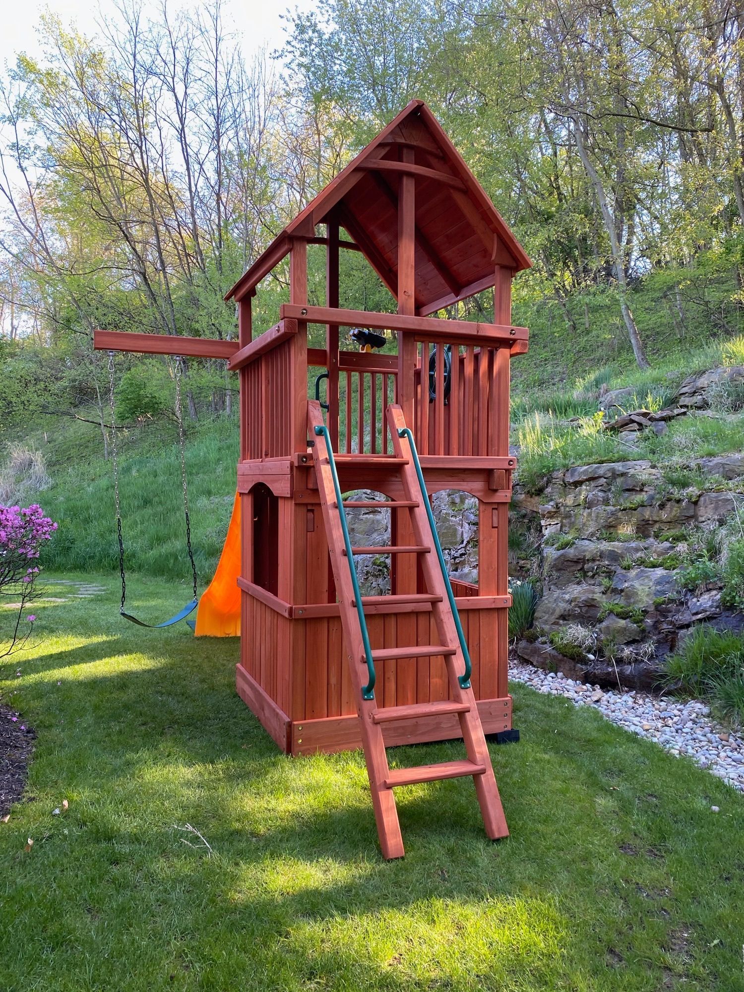 Red wooden backyard playset with slide and ladder on a grassy slope, near stone landscaping and trees