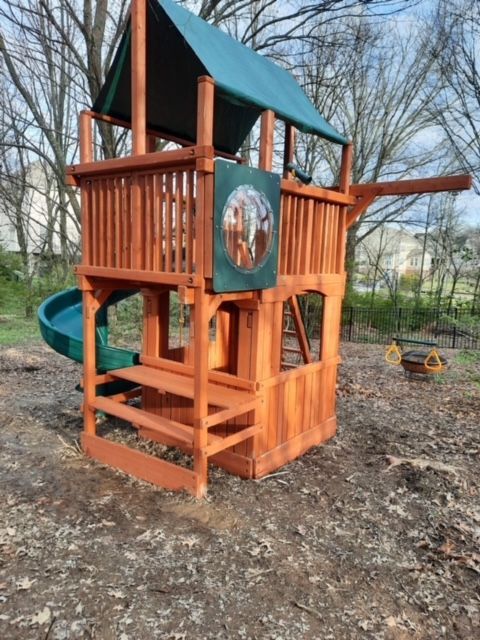 Wooden backyard playset with slide, green roof, and animal-themed panel on a dirt yard.