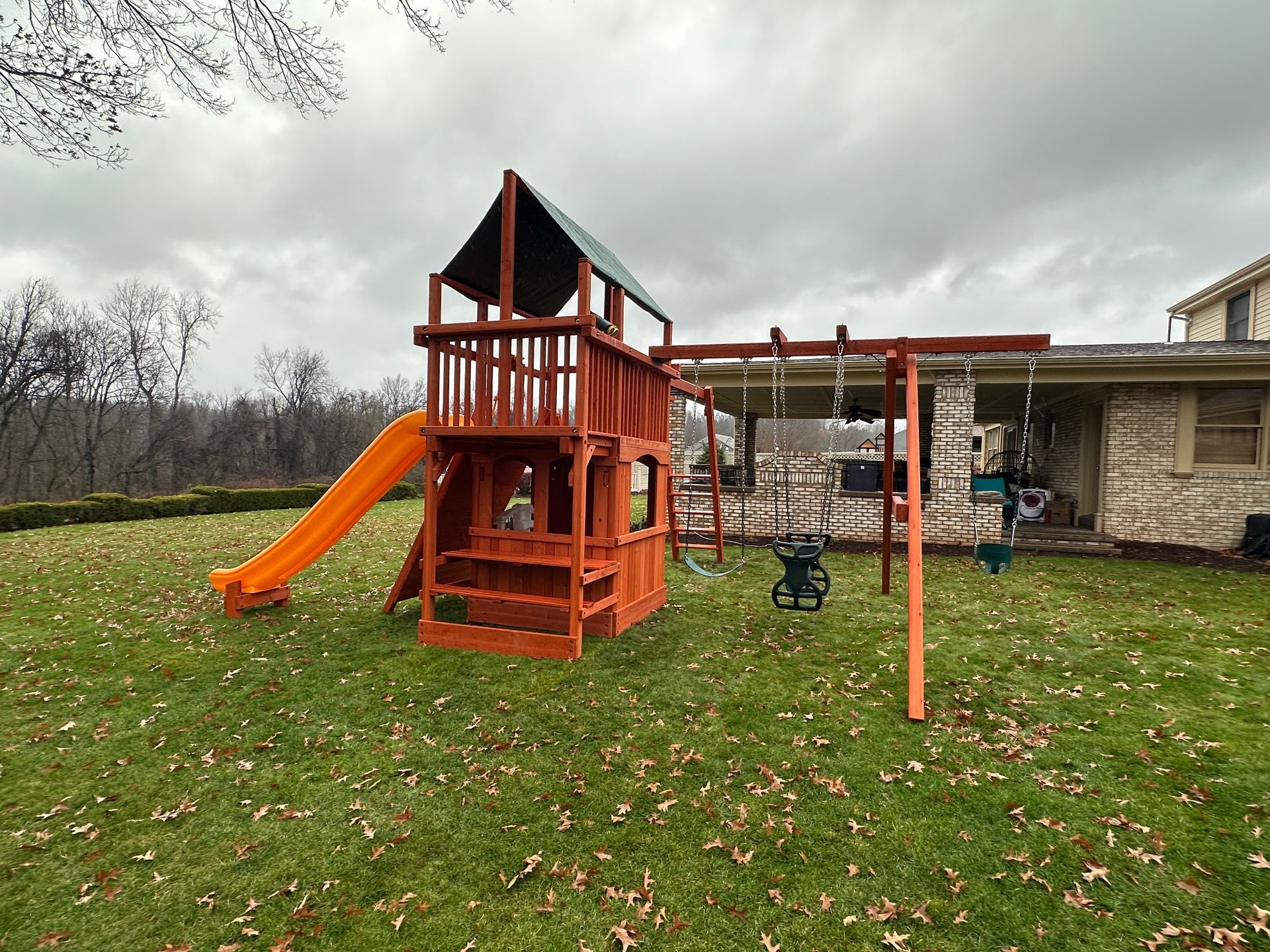 Backyard playset with orange slide, swings, and climbing tower on a grassy lawn under cloudy sky