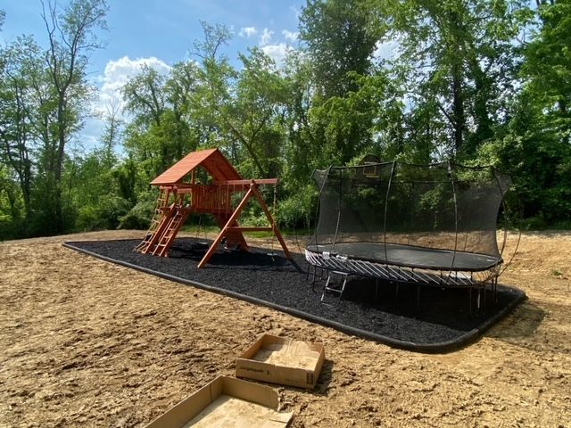 Children’s playground with red climbing tower and black trampoline on a wood-chip surface, surrounded by trees