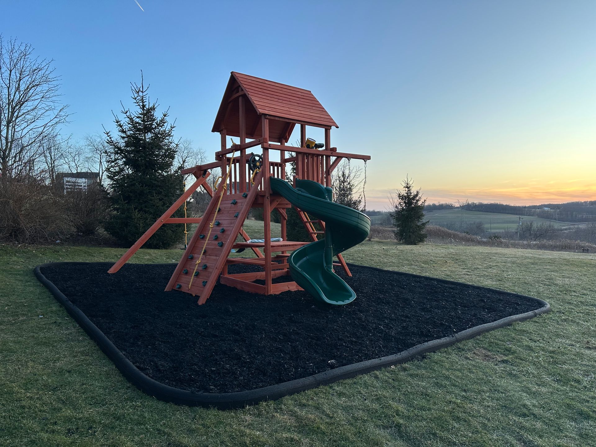 Wooden playground set with slides on a black mulch pad at sunset in an open field