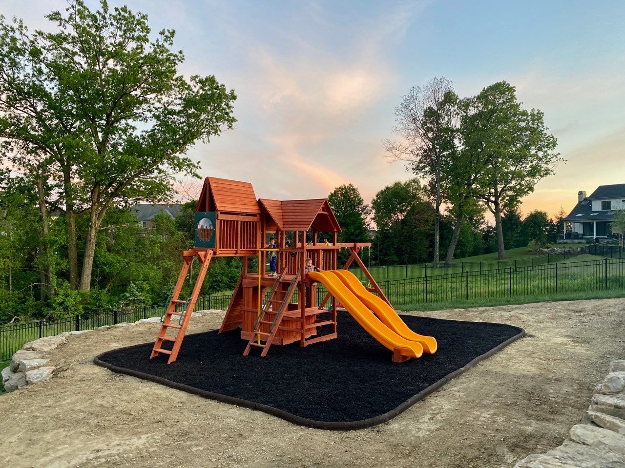 Wooden playground set with orange slides on a black mulch pad in a grassy park at sunset