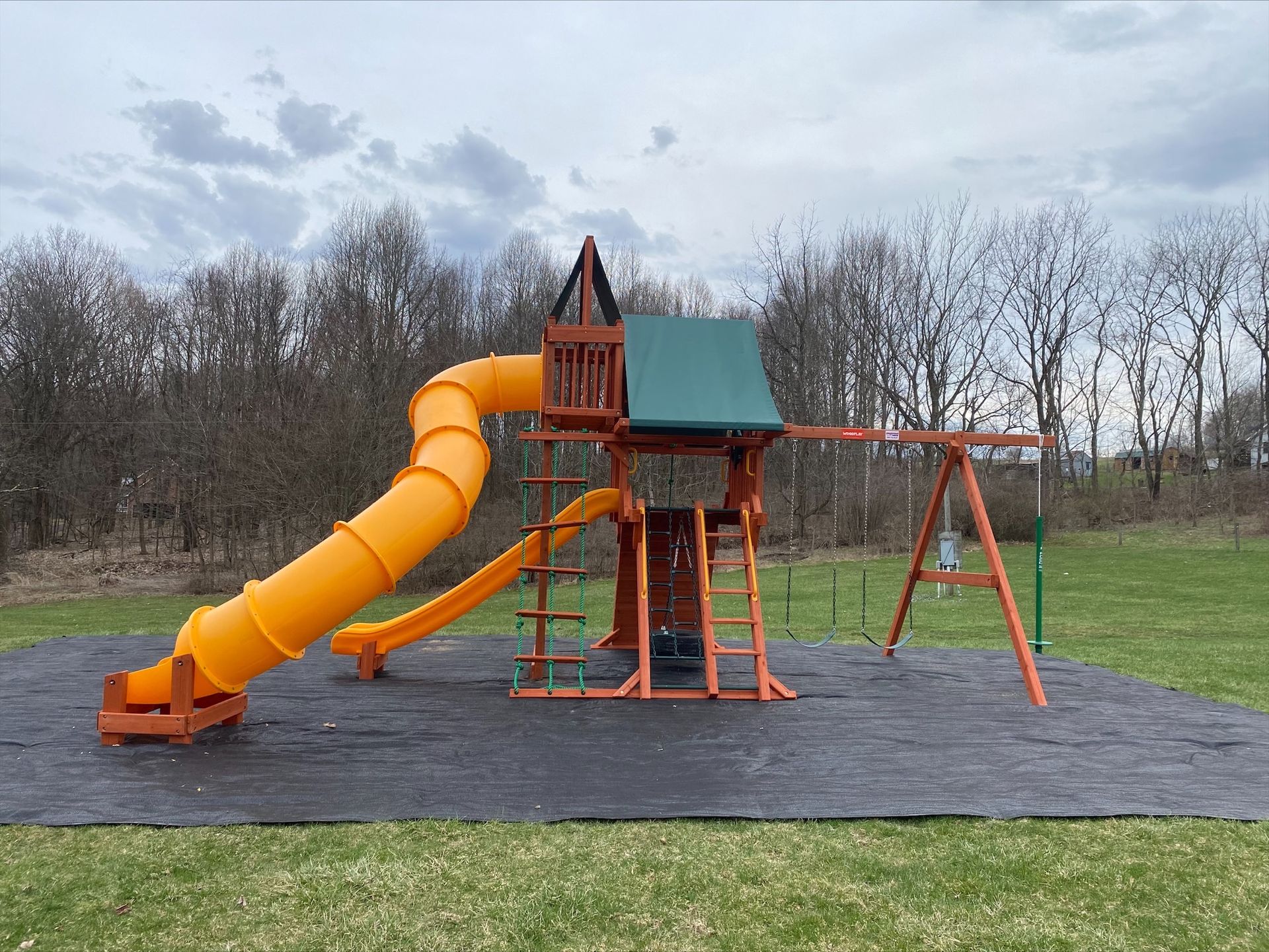 Orange playground slide and climbing structure on a rubber mat in a grassy park.