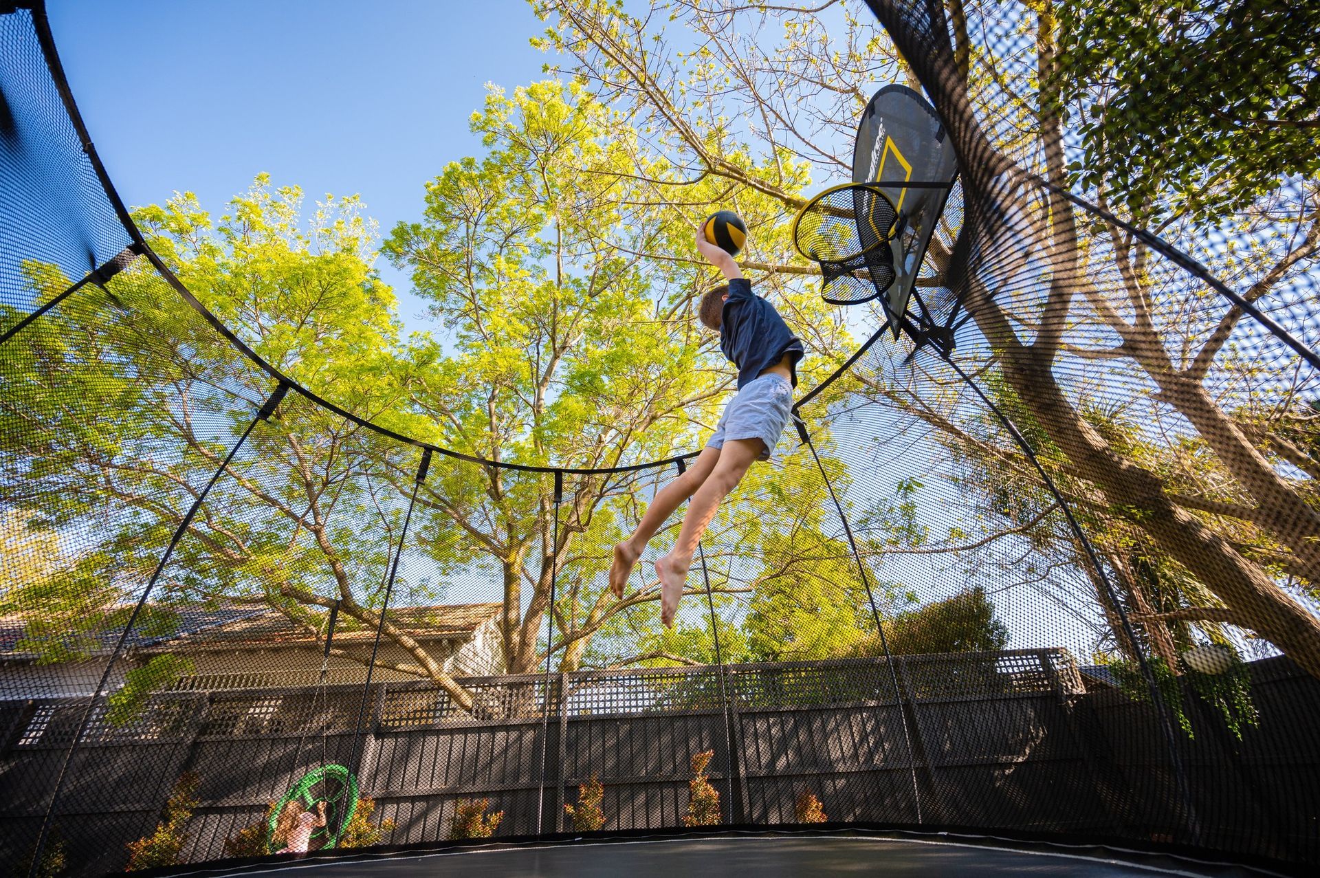 Person bouncing on an outdoor trampoline enclosed by netting, reaching toward a basketball hoop under sunny trees