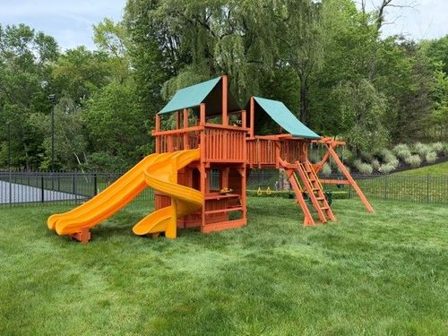 Colorful wooden playground set with slides and climbing ramps on a grassy lawn surrounded by trees.