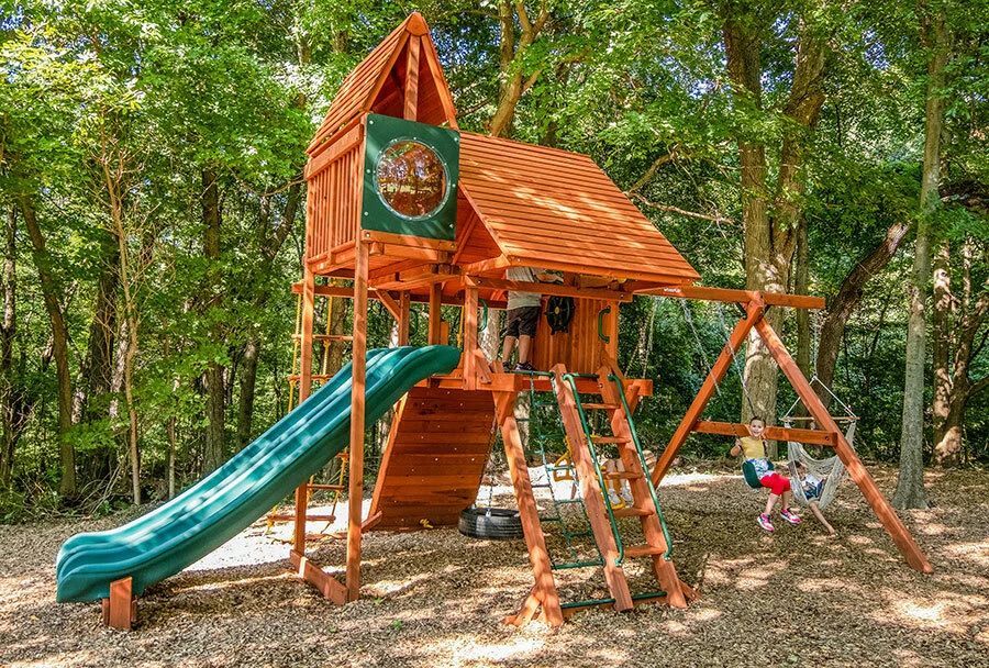 Wooden playground fort with green slide and swings in a wooded park, child playing nearby