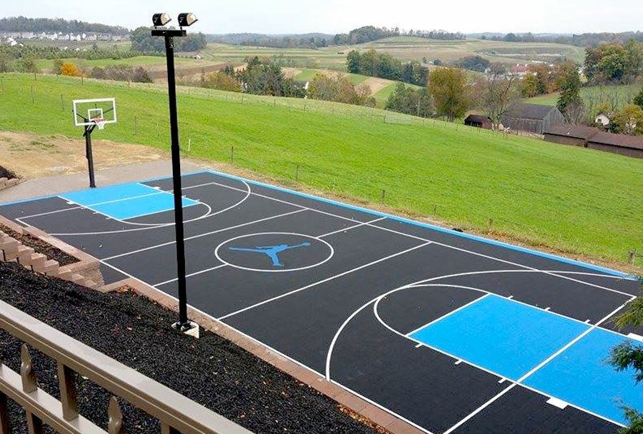 Outdoor basketball court with blue and black markings beside a grassy hillside and countryside view