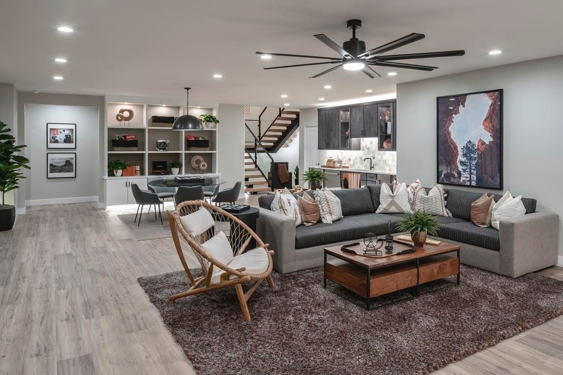 Modern open-concept living space with gray sofa, rug, armchair, and dining area. Kitchen and stairs visible in the background.