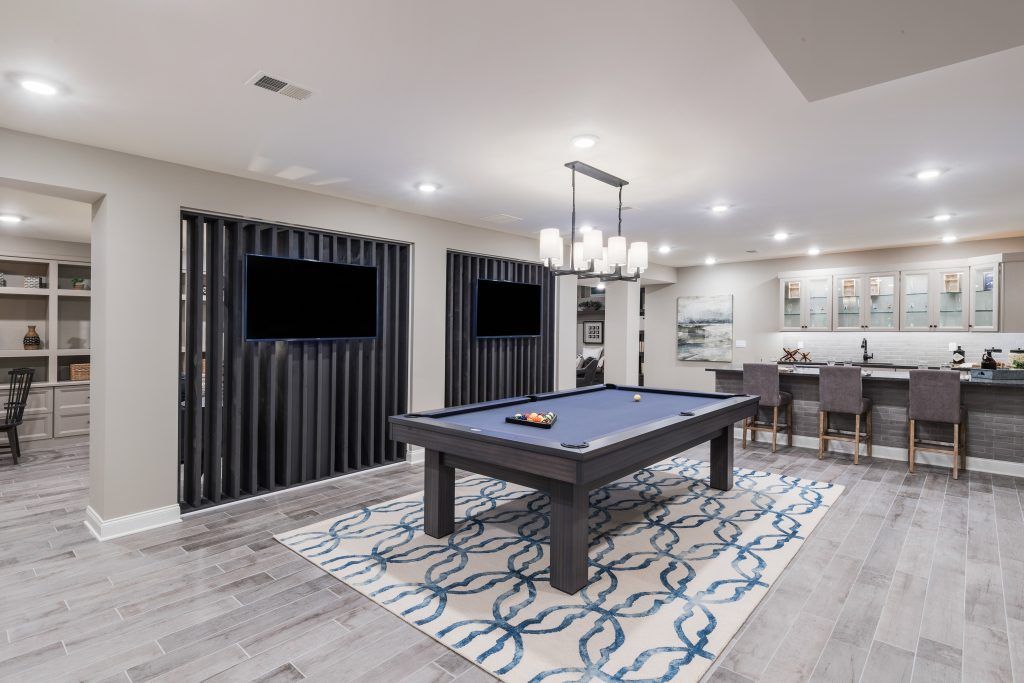 A finished basement with a pool table, bar, and TVs. Light gray floors, blue and gray accents, and modern decor.