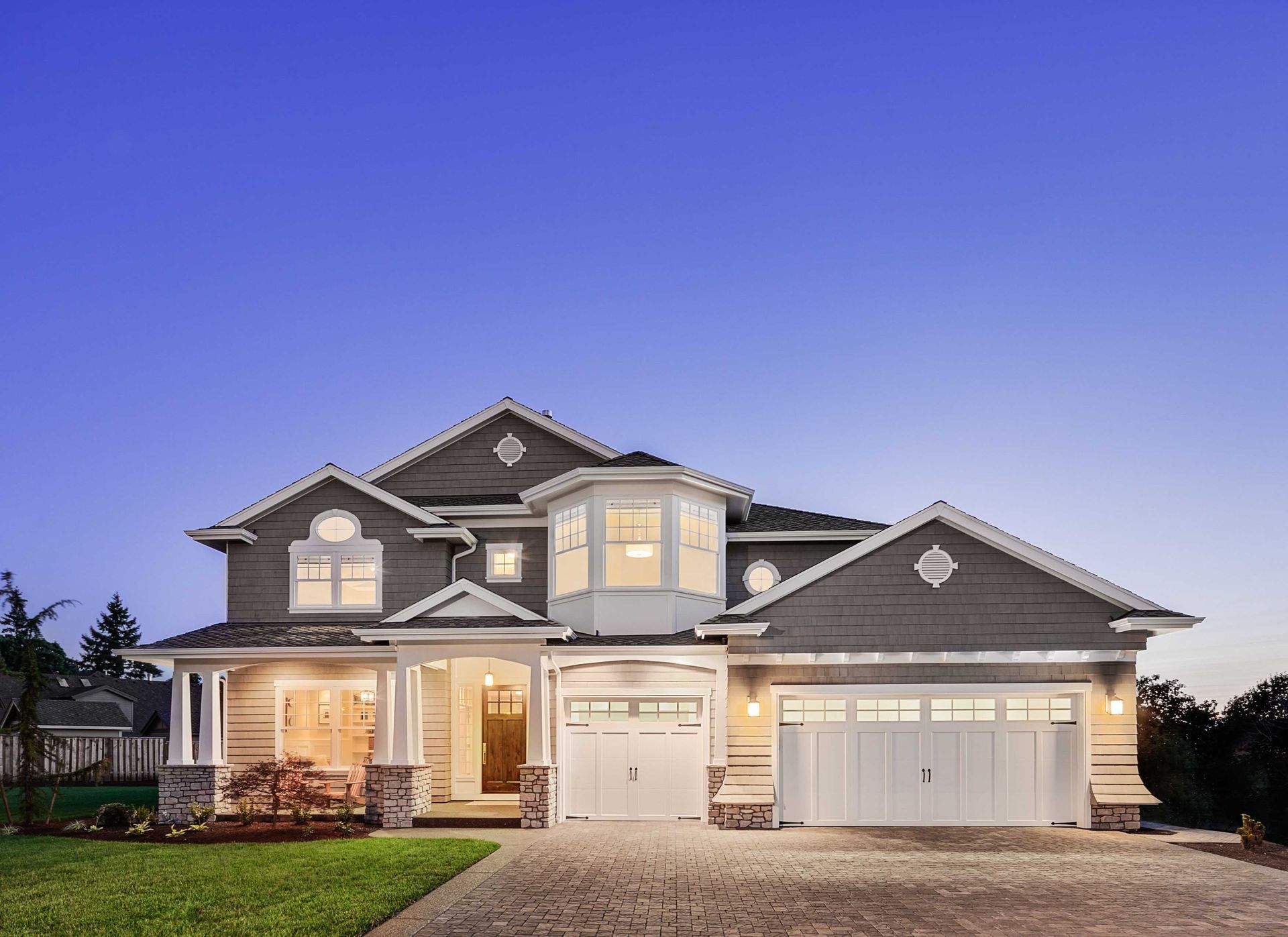 Two-story gray house with white trim, a bay window, two garage doors, and a stone driveway, at dusk.