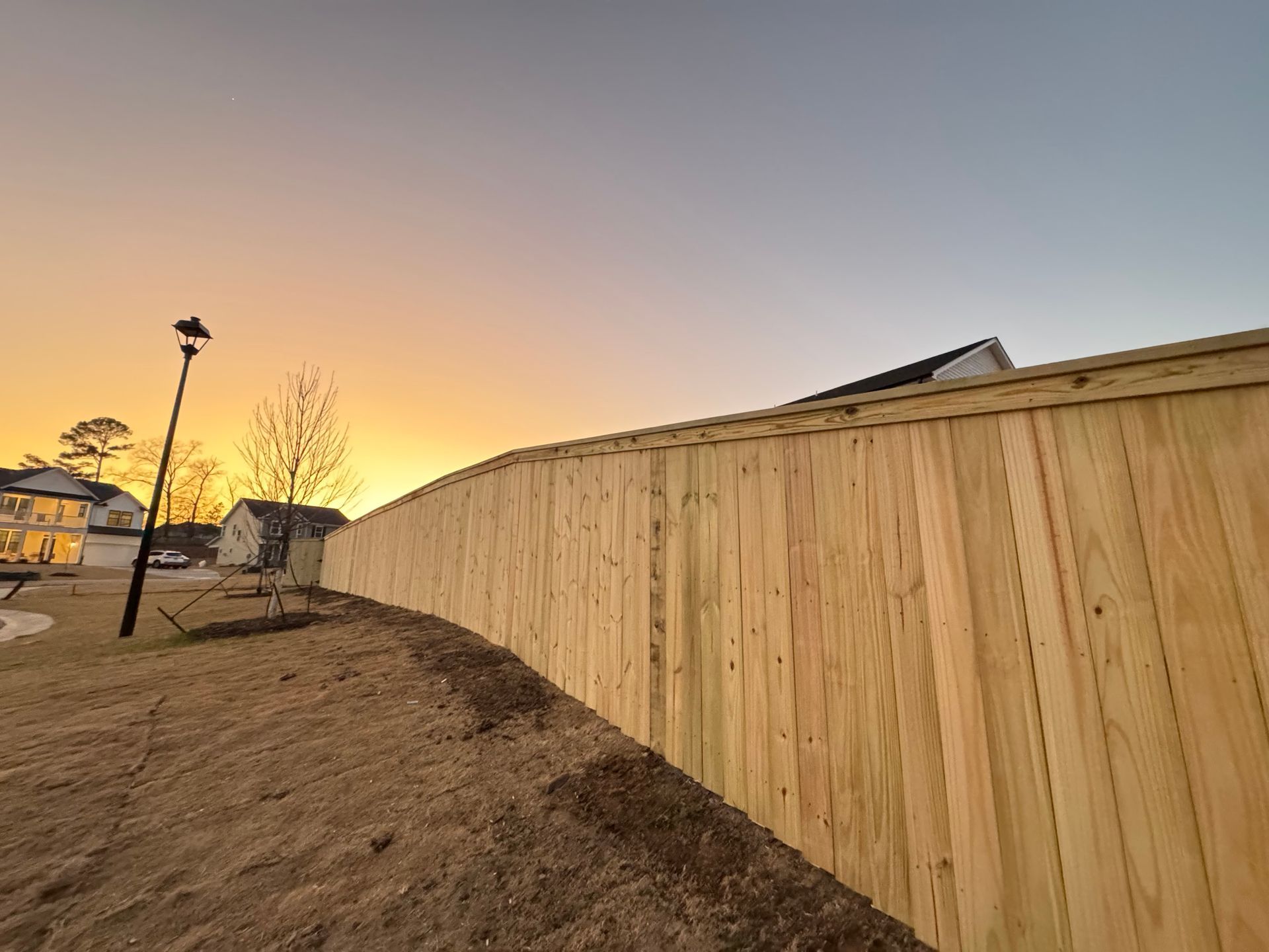 A wooden fence surrounds a dirt field with a sunset in the background.