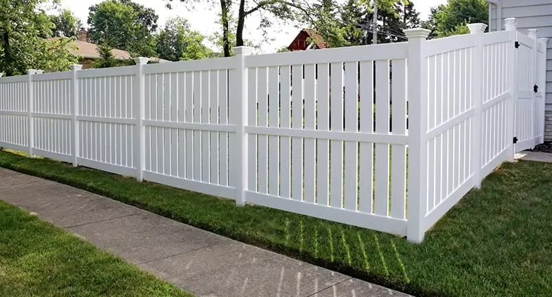 A white fence surrounds a lush green yard next to a sidewalk.