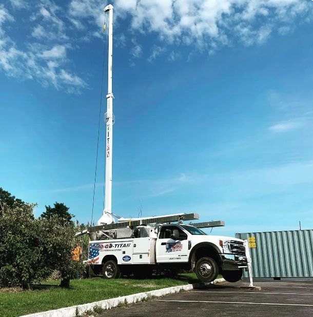 White utility truck with tall telescoping pole extended, parked on grass near a blue sky.
