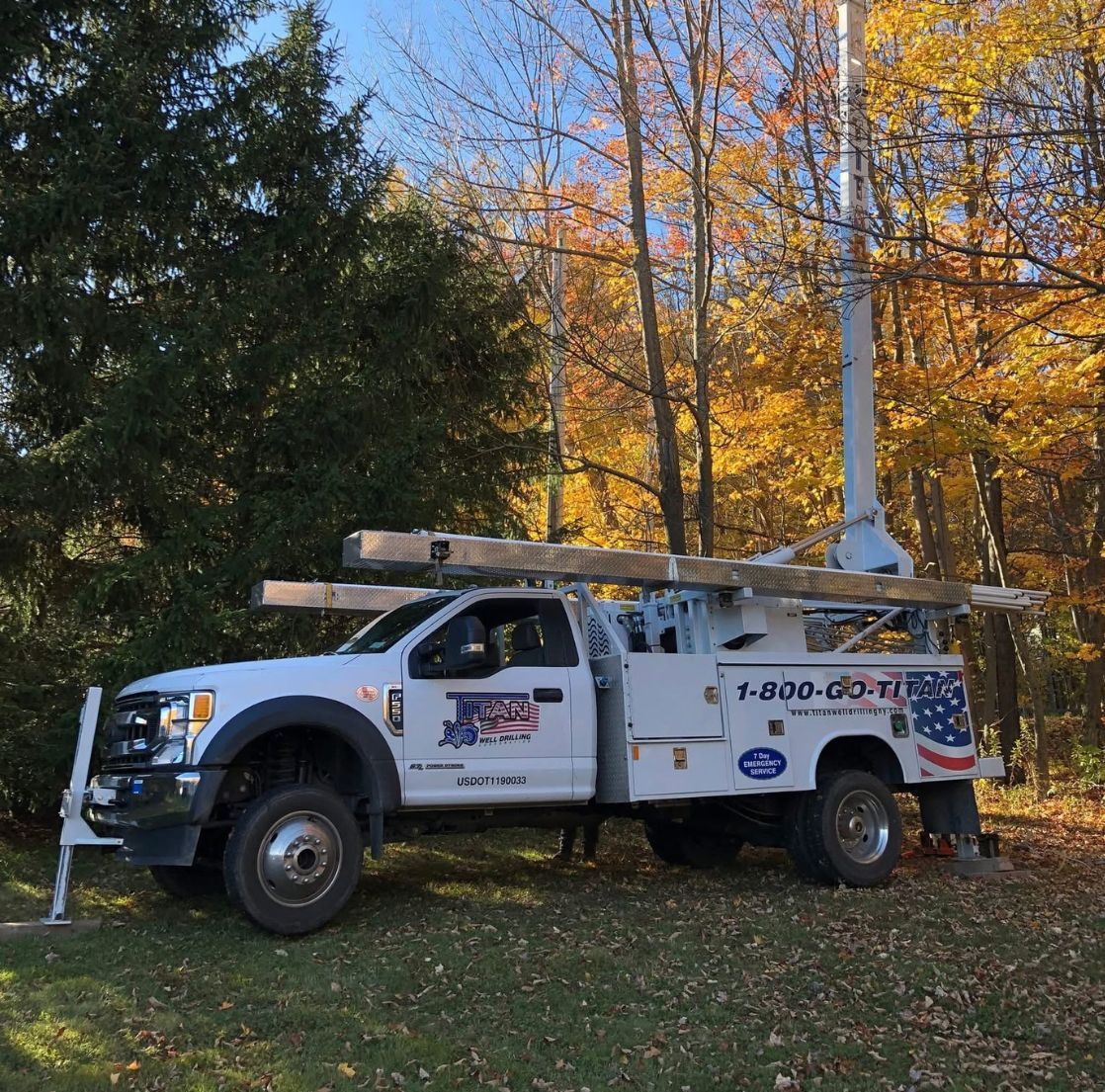 White work truck with a drilling rig parked on grass, autumn trees in the background.