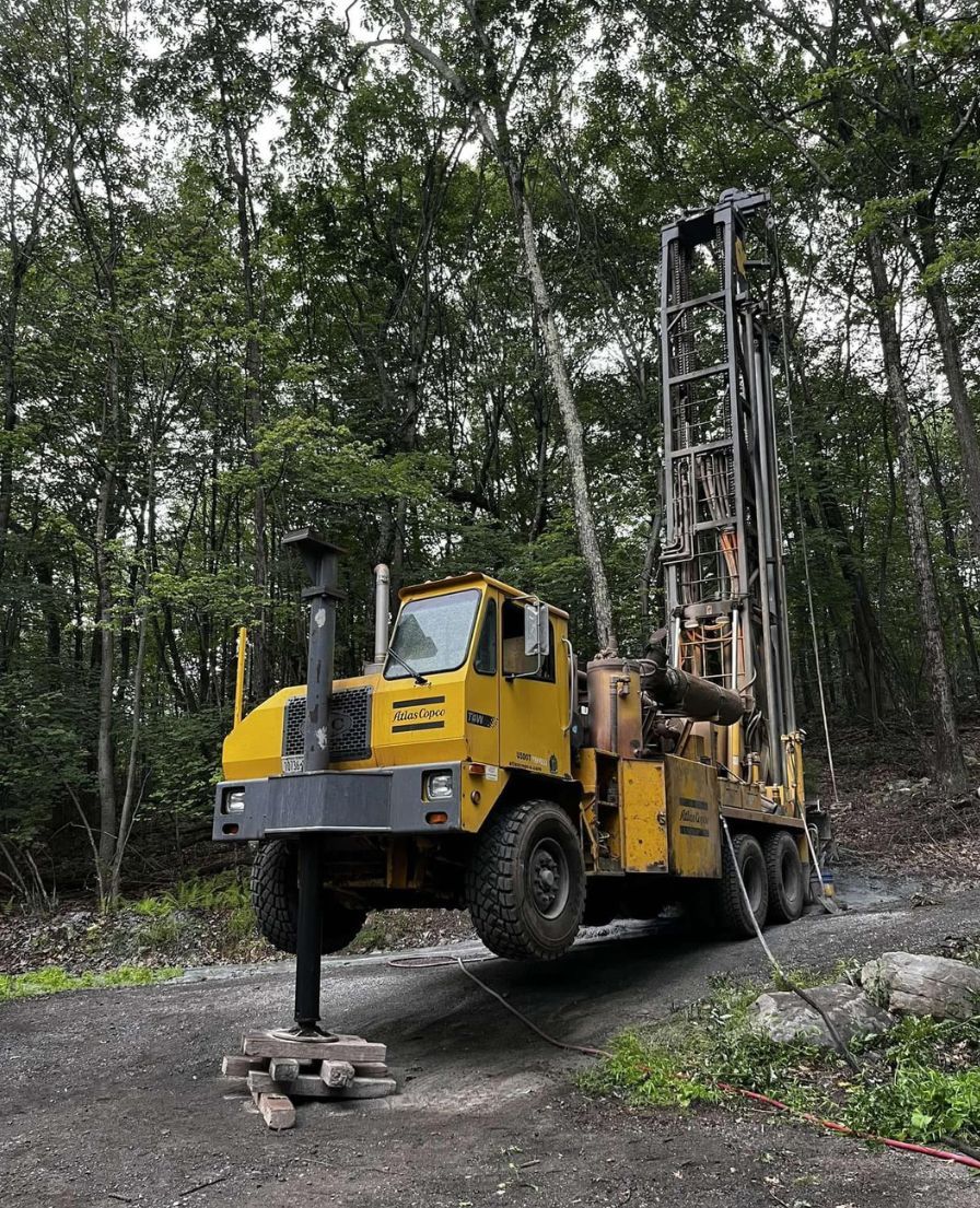 Yellow drilling rig on a gravel road in a wooded area, drilling into the ground.