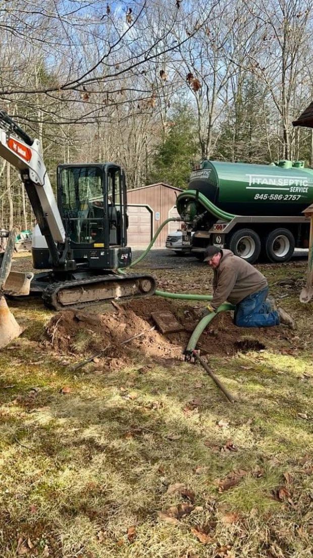 Man kneeling, connecting a green hose to a septic tank being excavated by a small excavator in a yard.