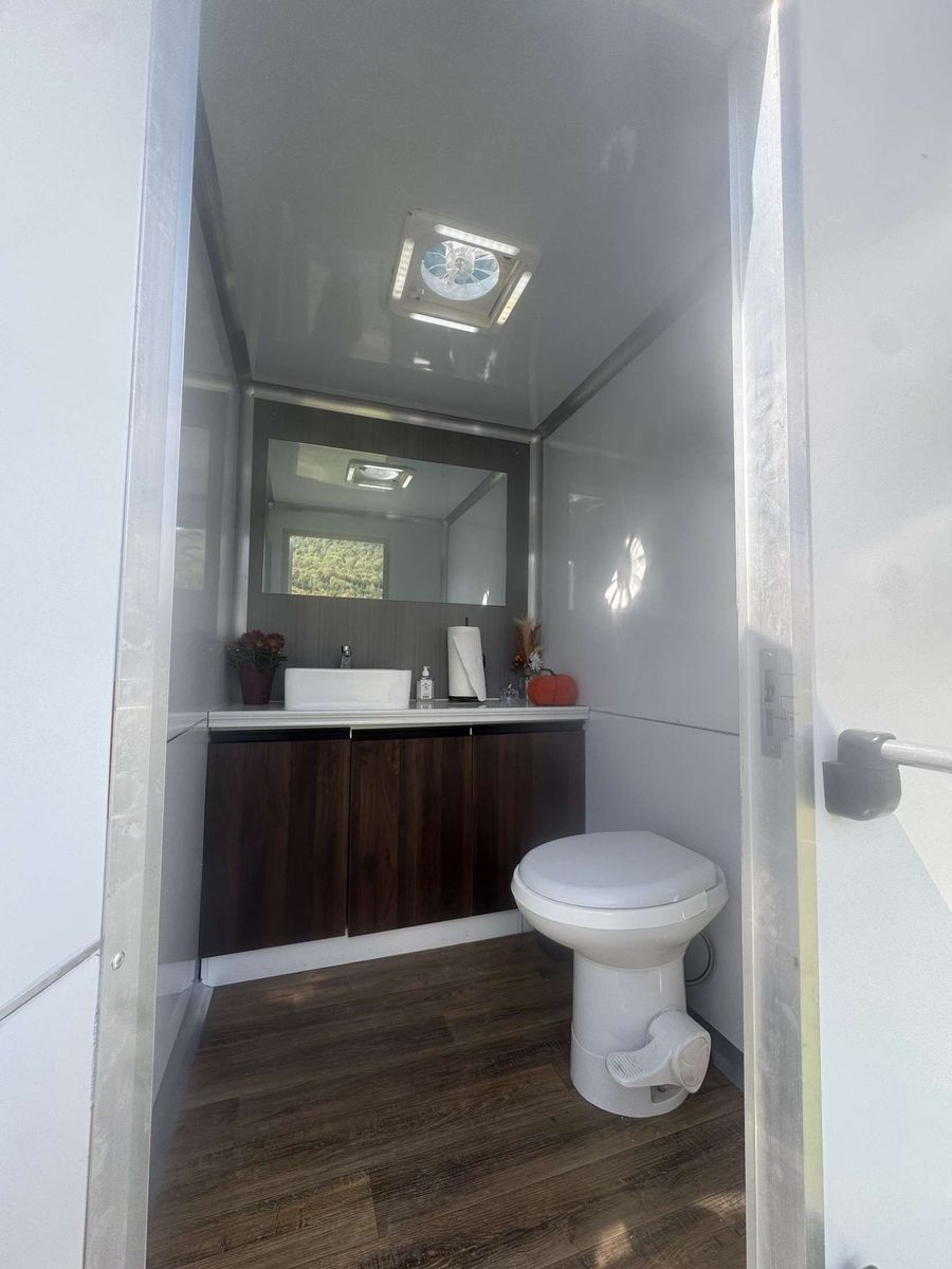 Interior of a luxury portable restroom. White walls, wooden floor, sink with mirror, and toilet.