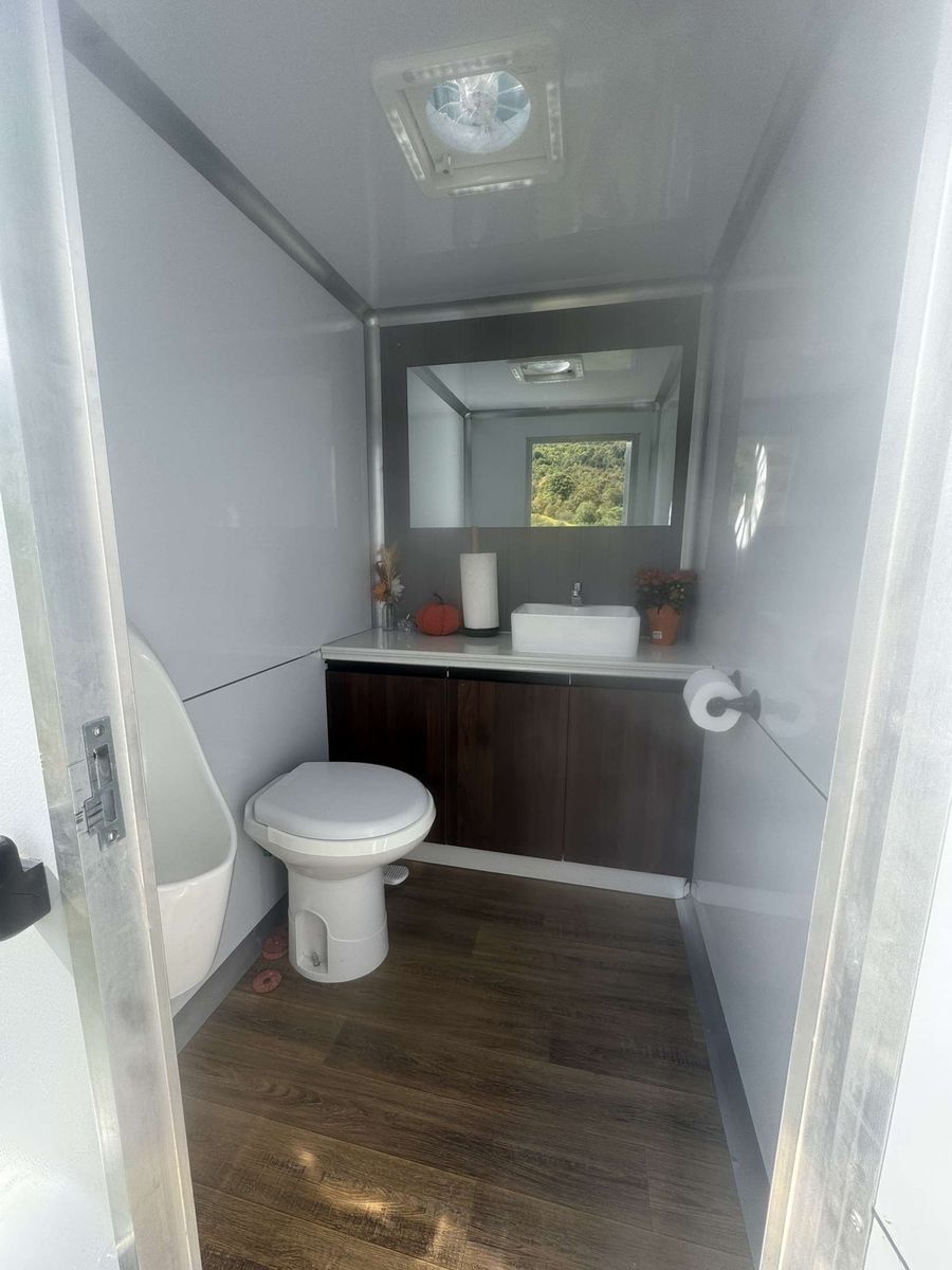 Interior of a portable restroom with a toilet, urinal, sink, and mirror. Brown cabinets and wood-look flooring.