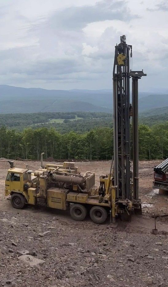Yellow drilling rig on a rocky hillside, tall tower extended. Mountains in the background.