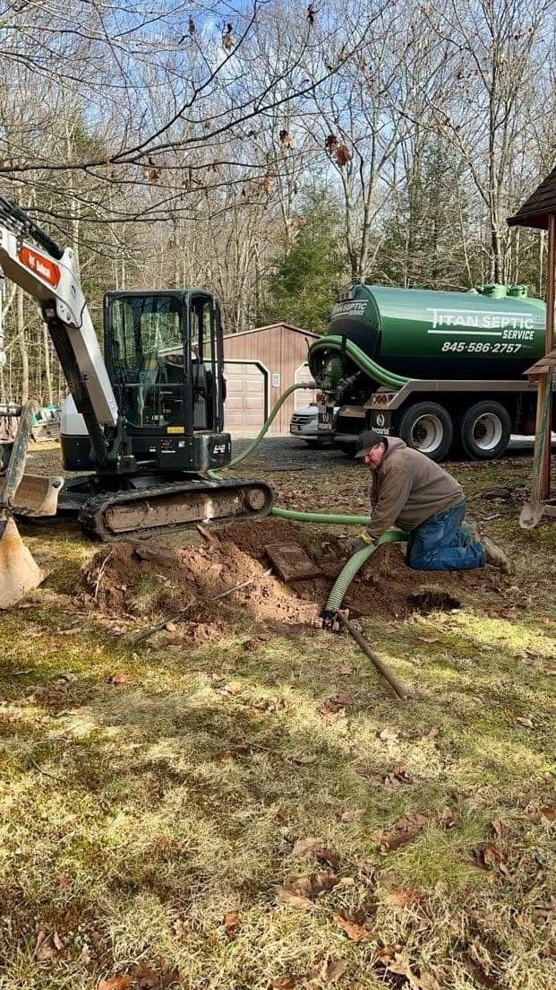 Person kneels, handling a hose near an excavation site. A mini excavator and green tank truck are present outdoors.
