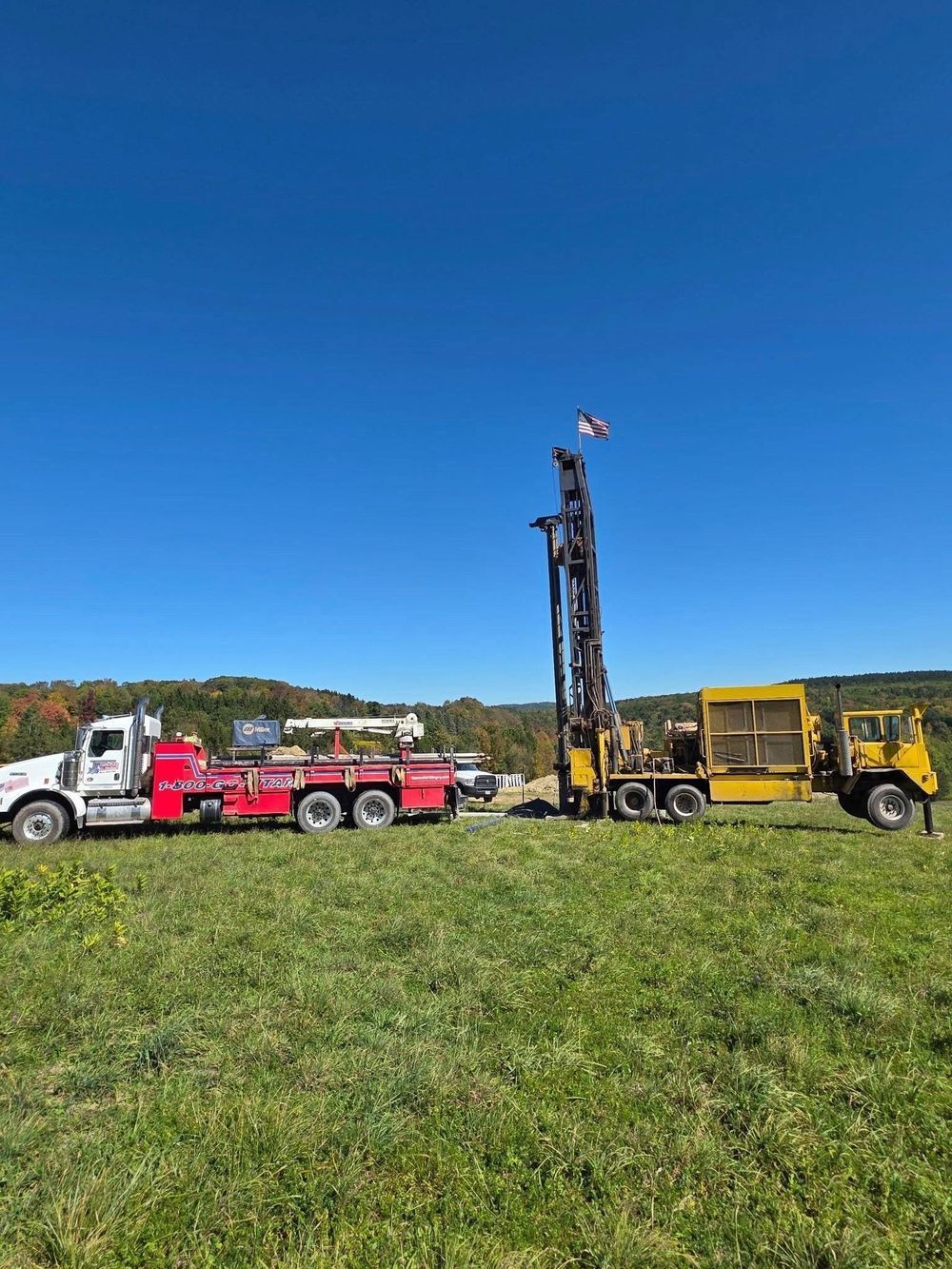 Red and white truck towing drilling rig on a green field under a blue sky.