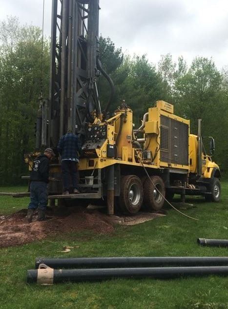Yellow drilling rig in a grassy area, two workers beside it. Black pipes on the ground.