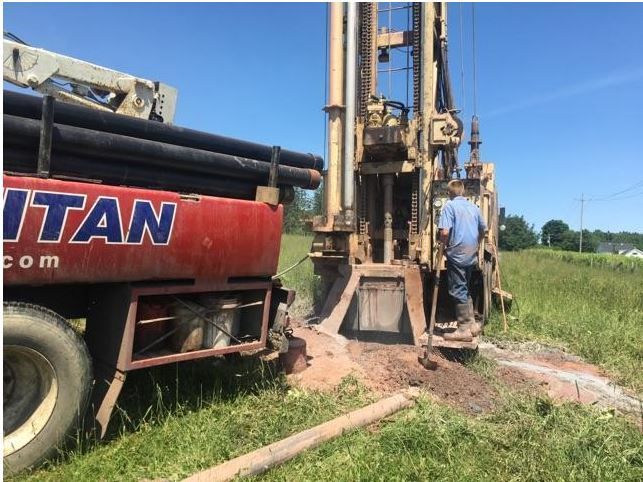 A well drilling rig is operating in a grassy field next to a red truck.