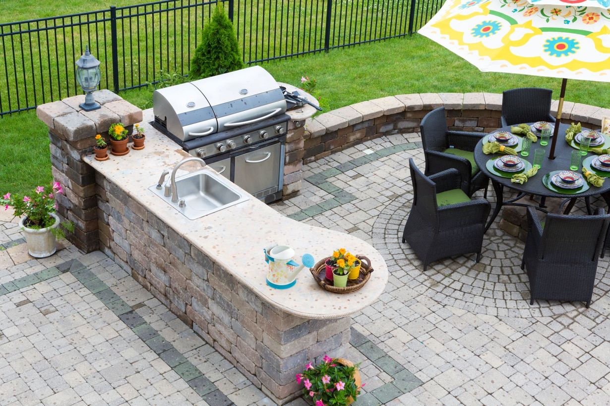 Outdoor kitchen with grill, sink, and countertop. Patio with dining table, chairs, and umbrella. Green grass, brick and black fence.