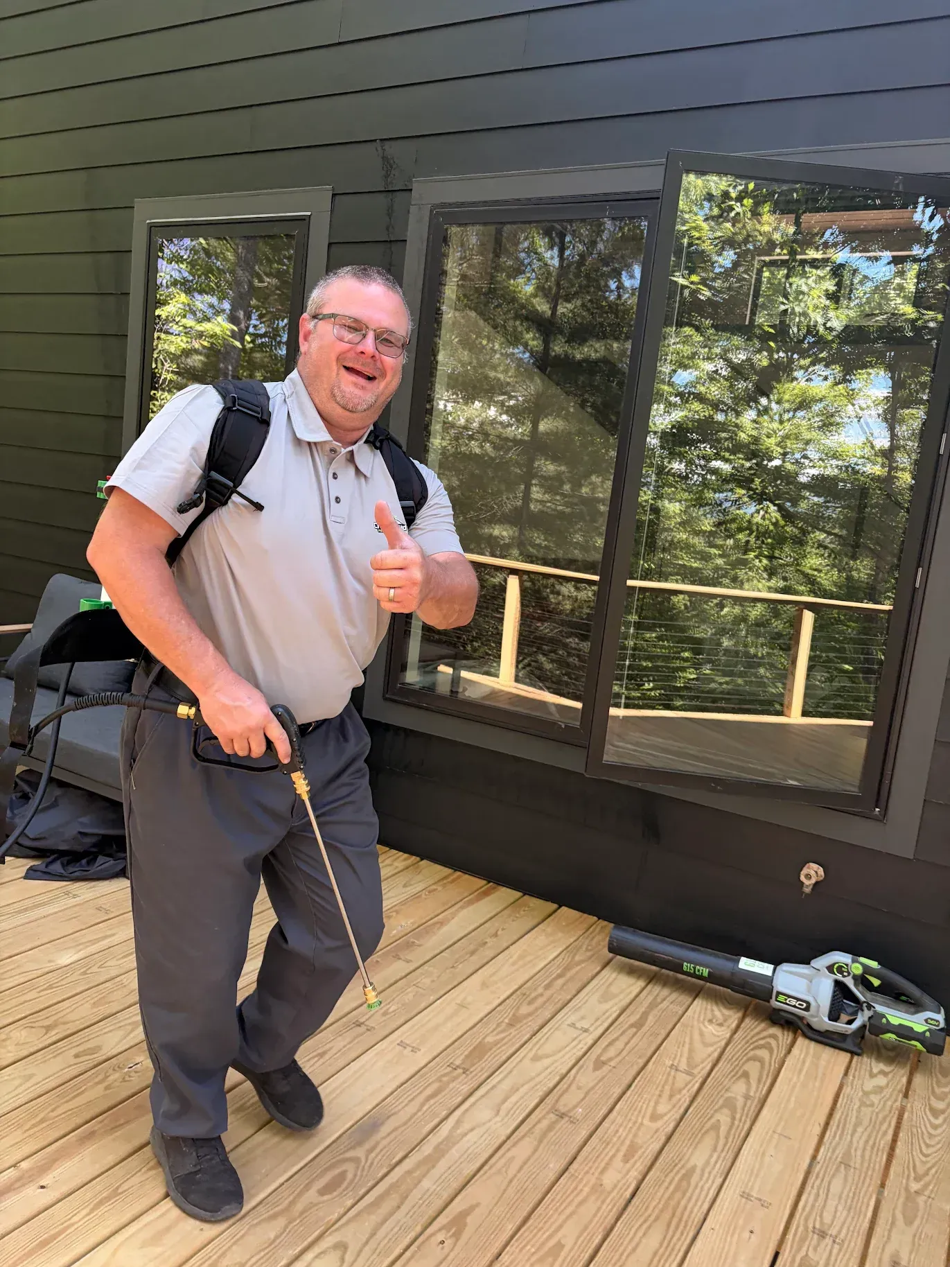 Man standing on a wooden deck by a house, smiling and giving a thumbs-up with a backpack on.
