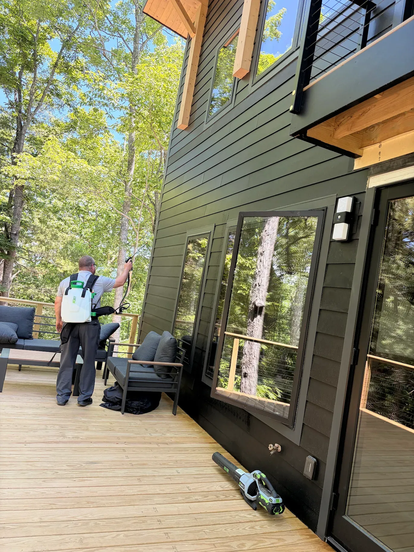 Two people standing on a wooden deck beside a black house with trees and lawn chairs nearby