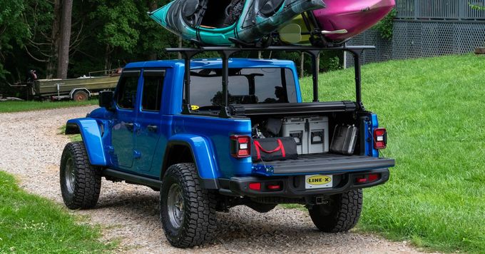 A bright blue Jeep Gladiator truck parked on a gravel driveway with two kayaks secured to a black bed rack.