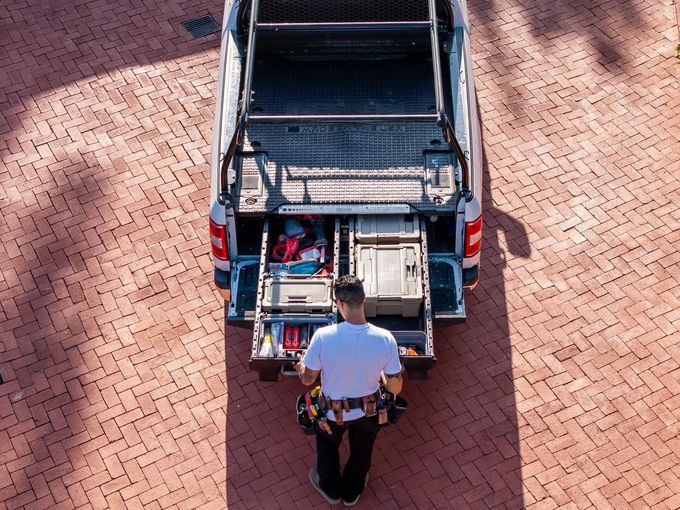 A person in a white t-shirt and tool belt accessing drawers in the bed of a truck parked on a brick driveway.