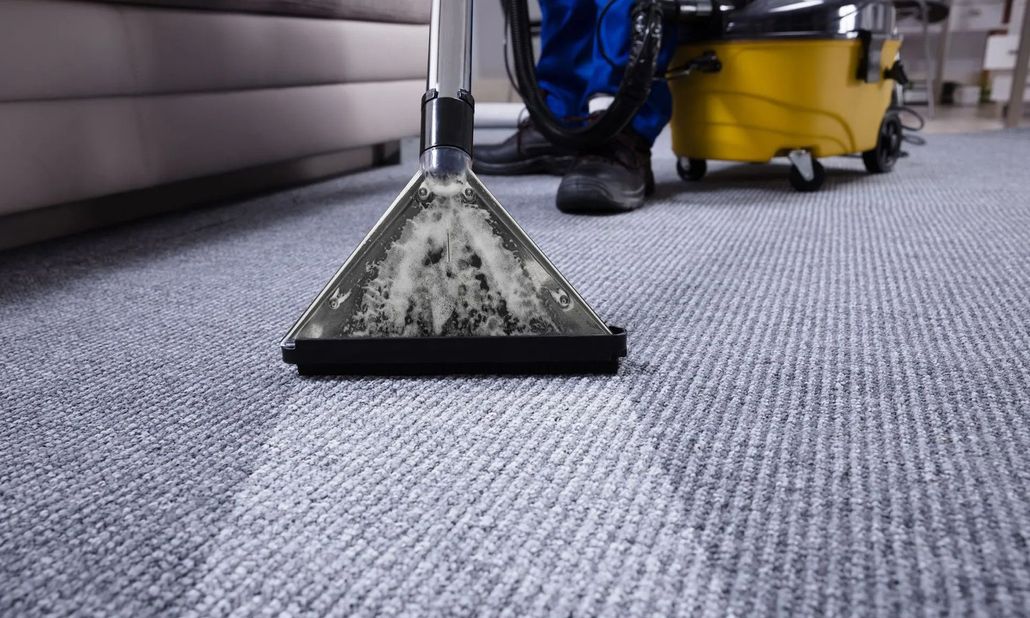 Carpet cleaning machine vacuuming a gray office carpet, with a person and yellow equipment in the background