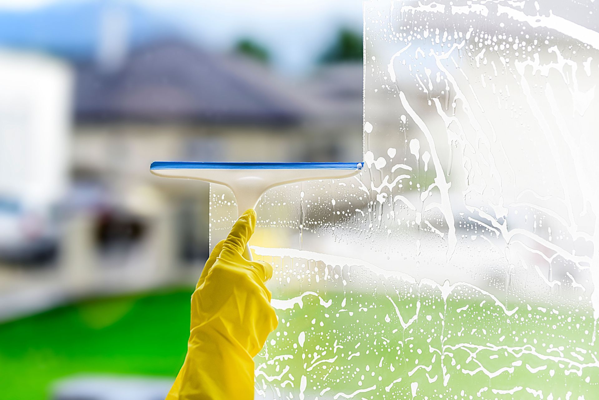 Gloved hand wiping a window with a squeegee, water spraying against a blurred house backdrop window cleaning