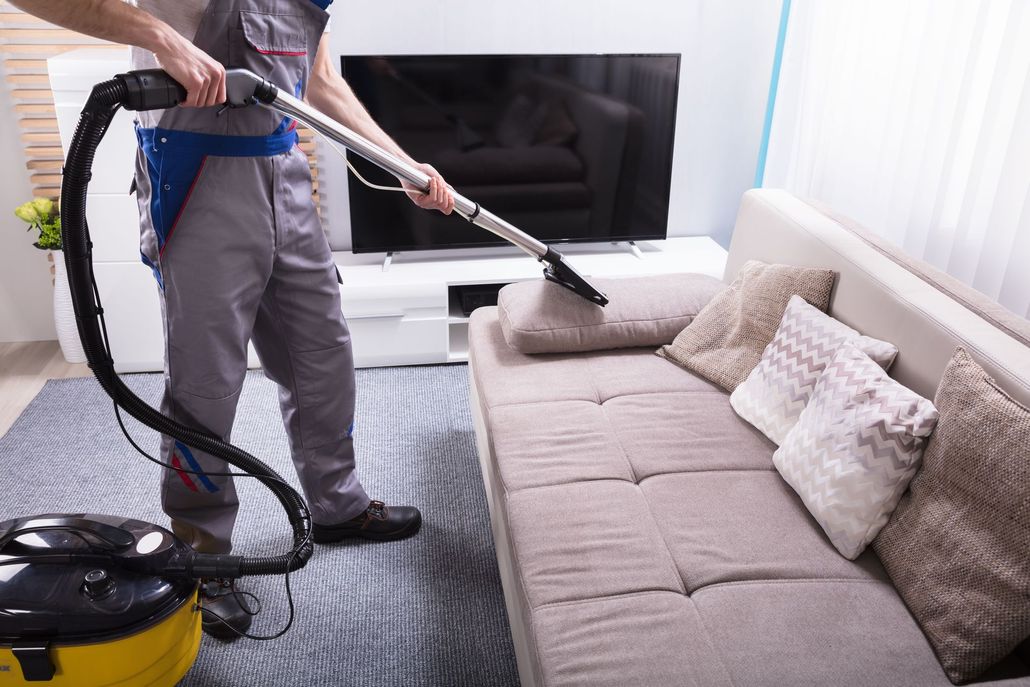 Person vacuuming a beige couch in a bright living room with a canister vacuum