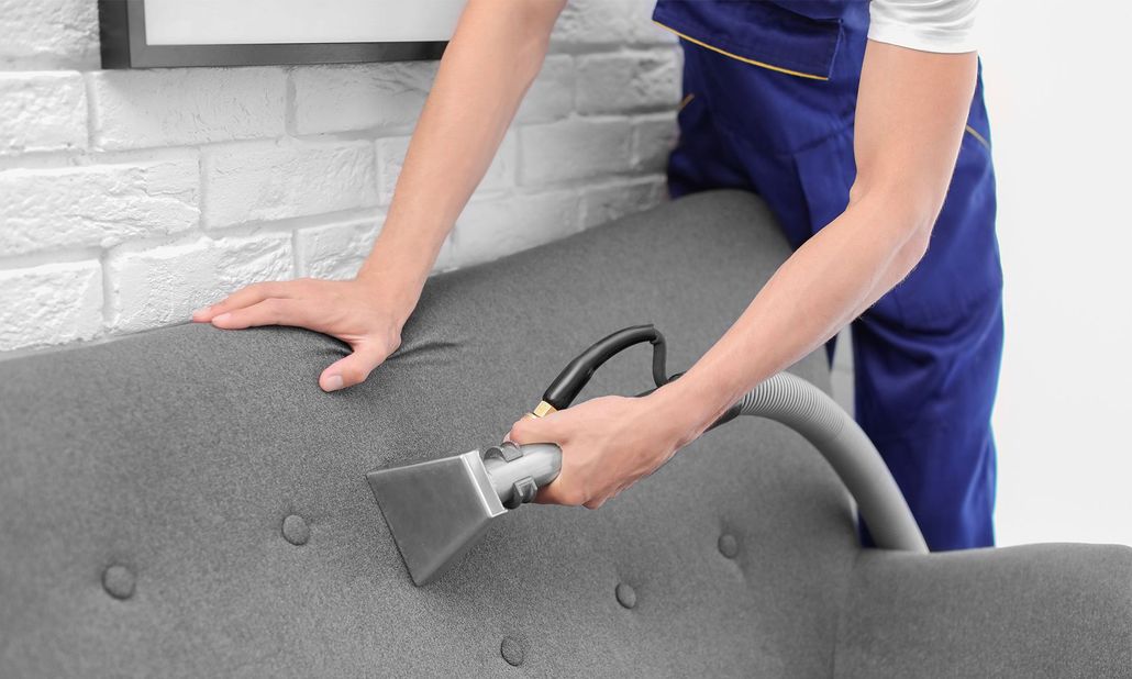 Person vacuuming a gray couch with a handheld upholstery cleaner in a bright room