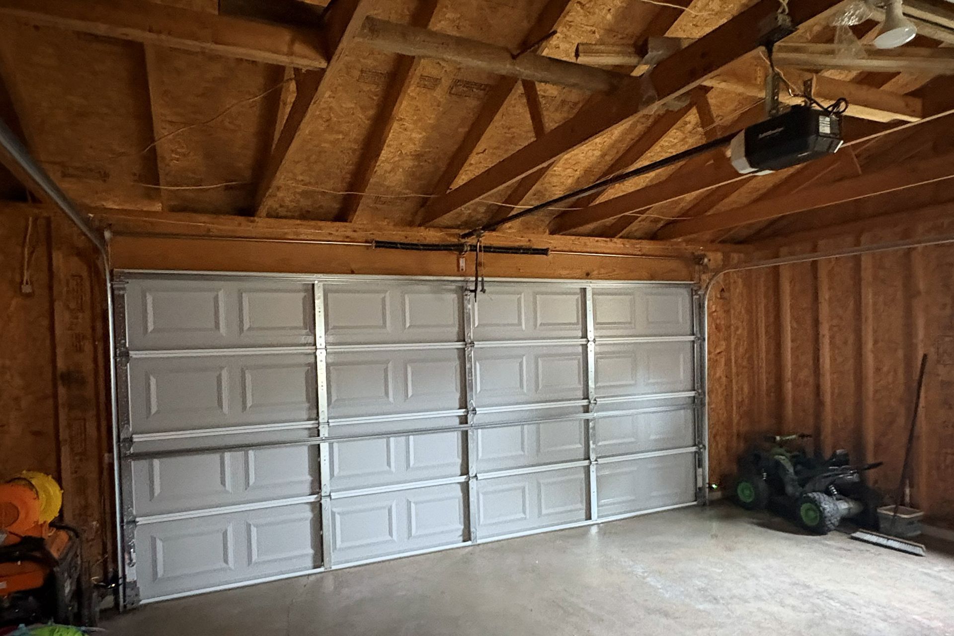 Garage interior with a partially open white garage door. A lawnmower and tools are visible.