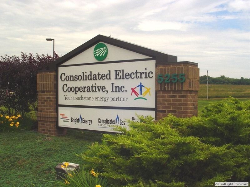 Sign for Consolidated Electric Cooperative Inc. with green logo and address 8250, brick and landscaping in front.