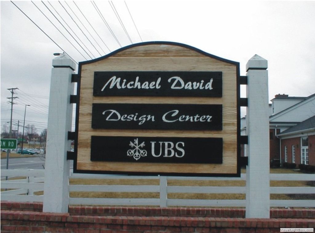 Sign for Michael David Design Center and UBS, with a white fence and brick base.