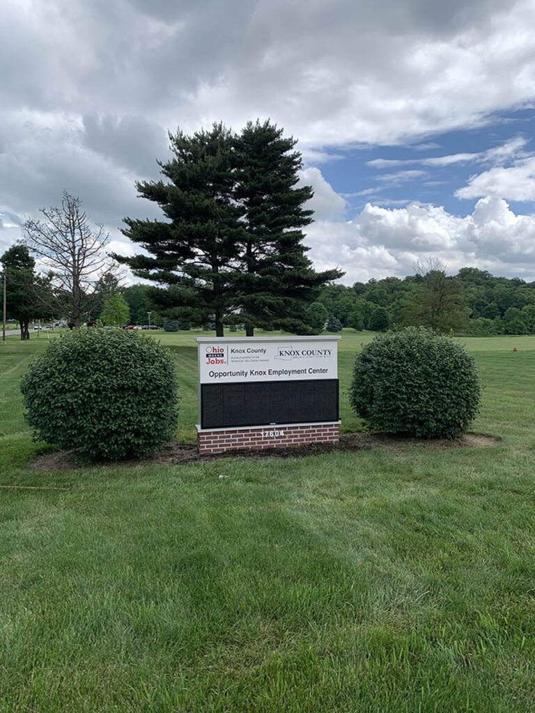 Sign with text, flanked by two round bushes, in a grassy area with trees and cloudy sky.