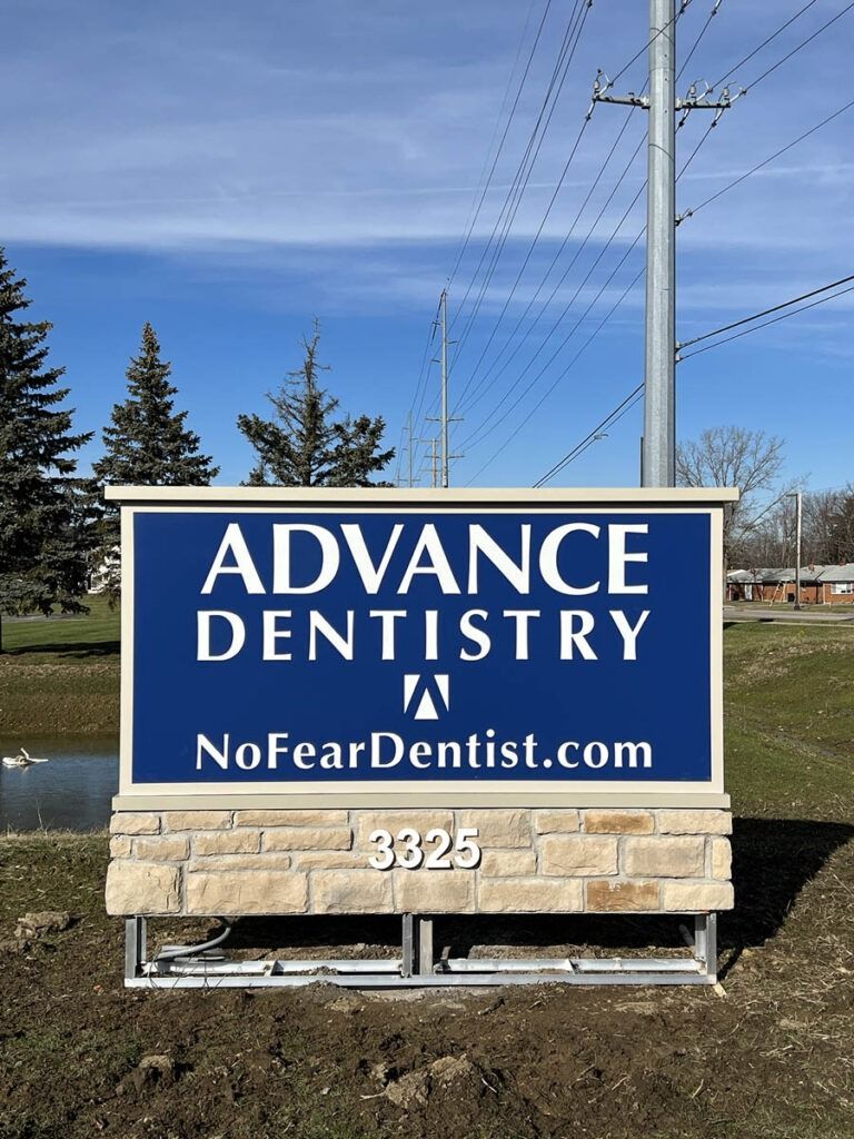 Sign for Advance Dentistry with blue and white text, a logo, and website address; set on a stone base, 3826 address visible.